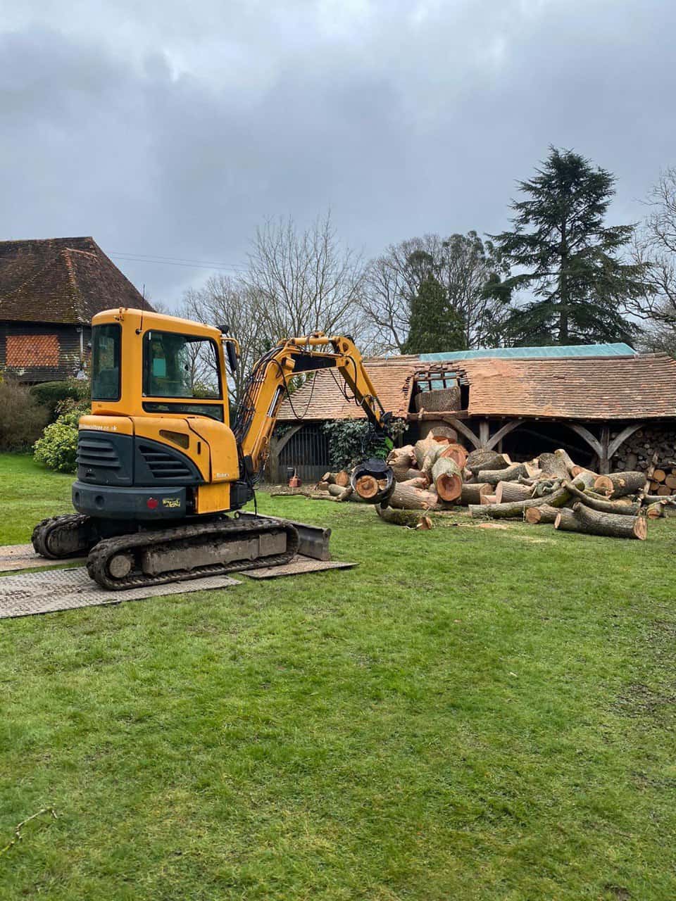 This is a photo of a tree which has grown through the roof of a barn that is being cut down and removed. There is a digger that is removing sections of the tree as well. Ilkeston Tree Surgeons